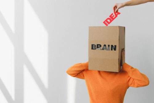 A person in an orange shirt holds a cardboard box labeled "BRAIN" over their head as a hand puts the word "IDEA" into the box.