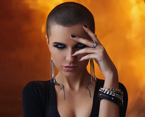 A person with dark, heavy eye make-up, super short hair, and black clothes stares intensely at the camera.