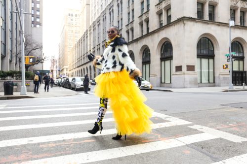 A person walks down a city street in a fluffy, bright yellow skirt, black and white leggings, a black and white sweater, and high heels.