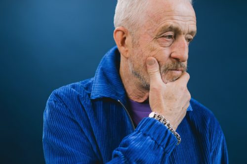 Older person in a blue jacket thinking with their hand on their chin.