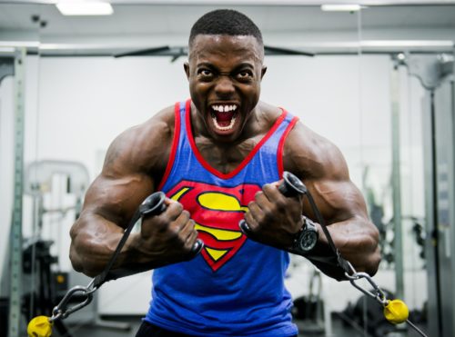 A person with big muscles yelling while working out in a Superman tank top.