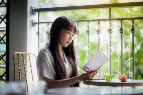Young woman reads a book at a small, round table beside a window.
