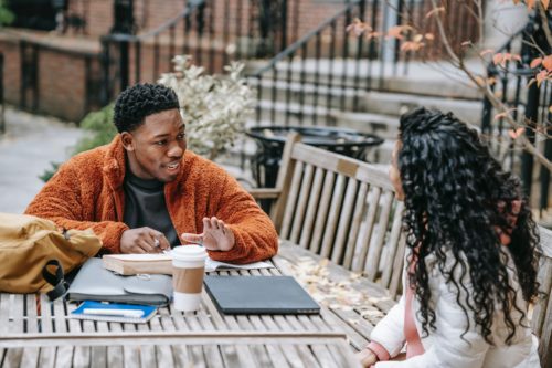 Young man and young woman talking over coffee, computer, and books at wood table outdoors.