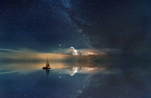 A sailboat on glassy water at sundown with clouds illuminated on the horizon and bright stars overhead.