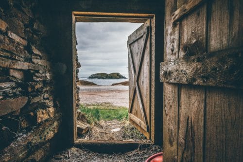 Open barn door looking out onto a beach scene with cloudy skies, sandy beach, and large, dark rocks in the ocean.
