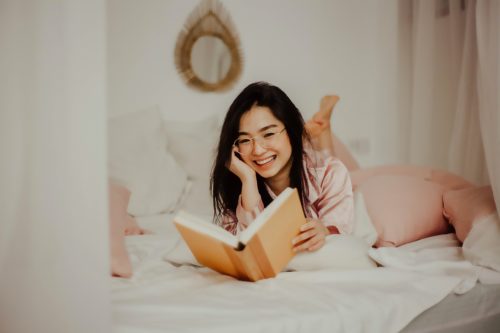 A smiling person with long hair and glasses lies on a bed reading a yellow book.