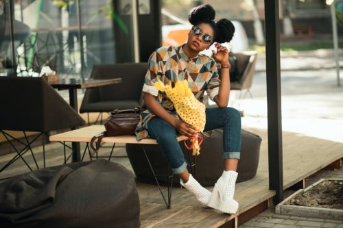 Person wearing round sunglasses, white platform shoes, jeans, and a button-up shirt with a geometric pattern sits outside a cafe holding a bouquet of yellow flowers.