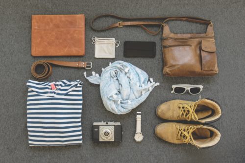 Items of clothing laid out in an organized pattern: striped shirt, scarf, camera, watch, belt, phone, sunglasses, boots, and purse.