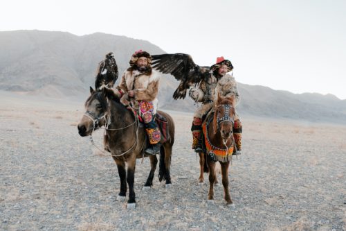 Two Mongolian falconers dressed in fur, hats, and colorful pants ride on horseback with their falcons.