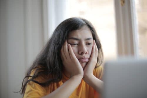 A bored-looking person holds their face in their hands as they stare at a laptop screen.