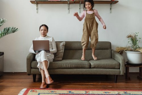 A woman sits on a gray couch writing on a laptop as her child jumps up and down beside her.