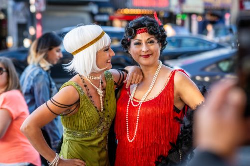 Two women dressed as flappers—one in a red dress and one in a green dress—smile together in an urban setting.