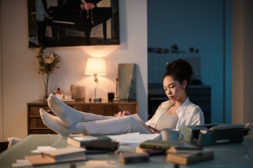 A person with a ponytail writes in a journal with their feet propped up on a table filled with books and notebooks.