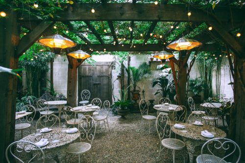 An empty cafe patio with small, round tables, soft lighting, a wooden back gate, and greenery overhead.