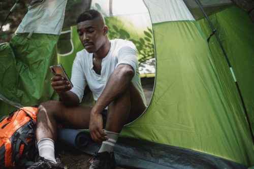 A man sits in the doorway of a green tent, looking at his phone.