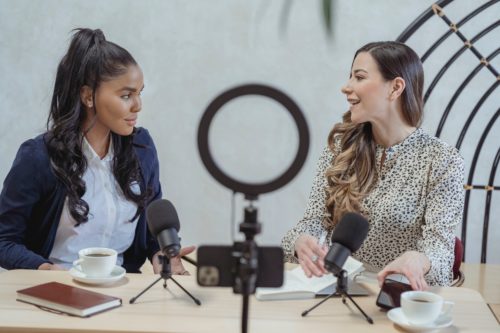 Two women sitting at a table behind microphones, doing a podcast interview.