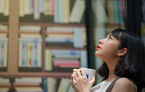 A person stands beside a book shelf, holding a mug and looking upward.