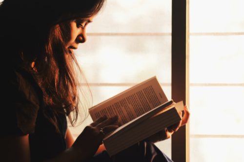 A person with long hair reading a book beside a window in a dark room.