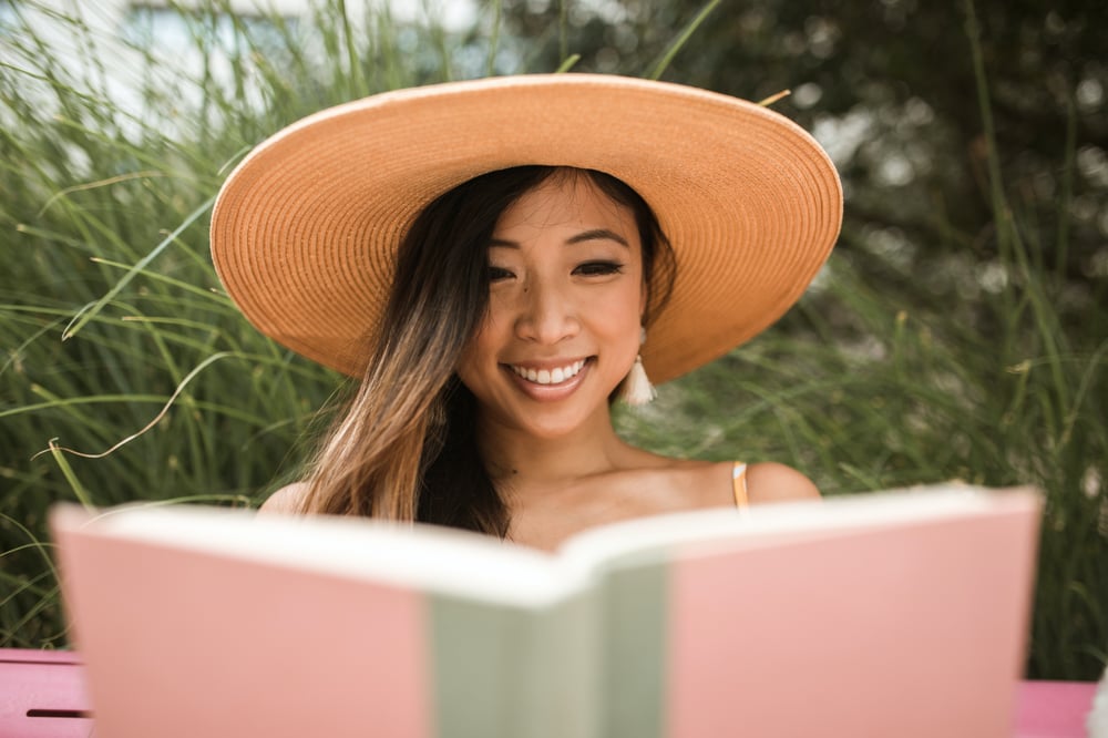 A woman in a wide-brimmed hat smiling as she reads a pink book.