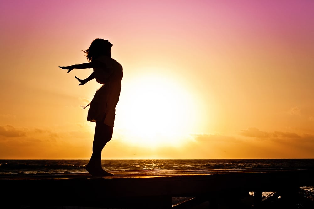 Profile of a person in a dress on a beach at sunset with their arms spread wide and their face towards the sky.