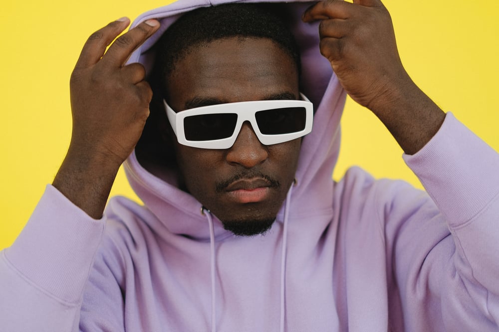 A person with a mustache and white sunglasses pulls the hood of a purple hoodie over their head.