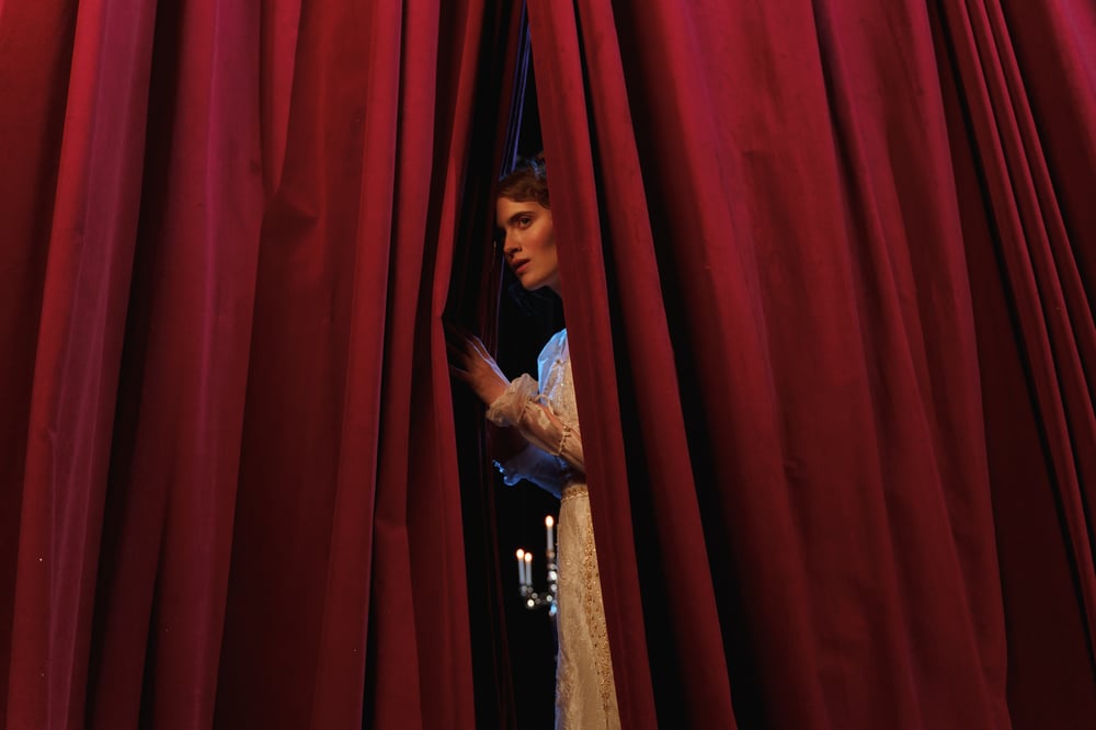 A person in a long, white Victorian gown peers through a gap in a red stage curtain.