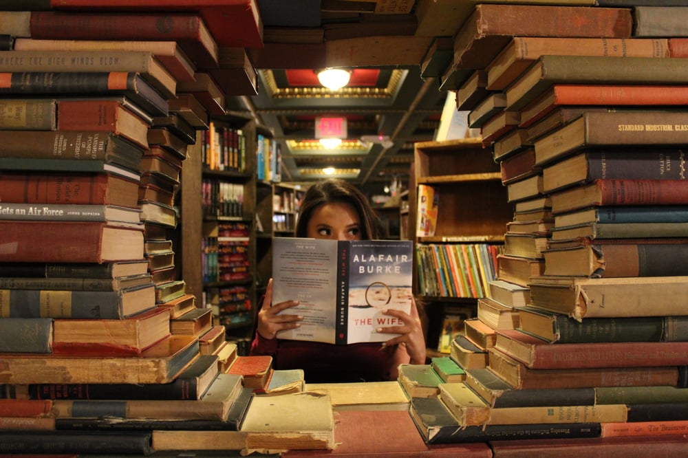 A person in a boostore looking through a round window made of books, holding the book The Wife by Alafair Burke in front of the lower half of their face.