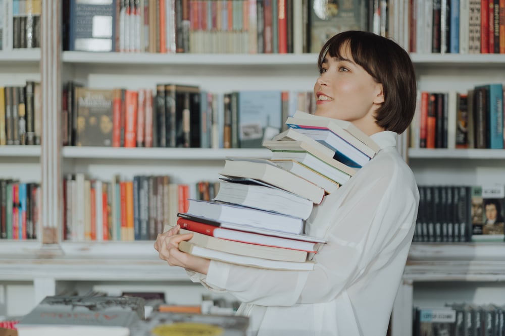 A person stands in front of a full bookshelf, holding a giant stack of books against their chest.