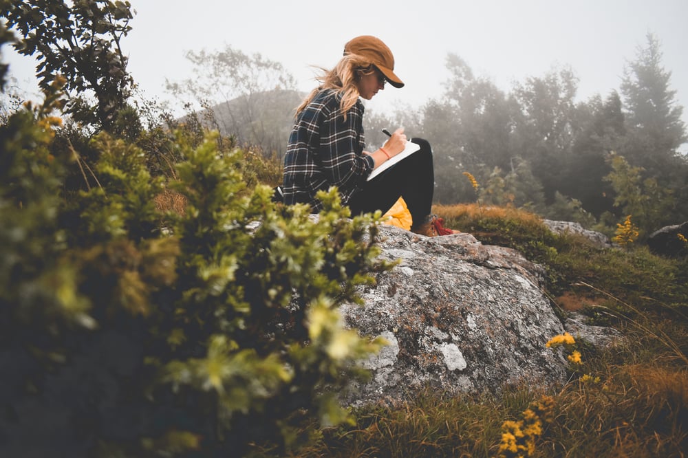 A person sitting outside on a rock writing in a notebook.