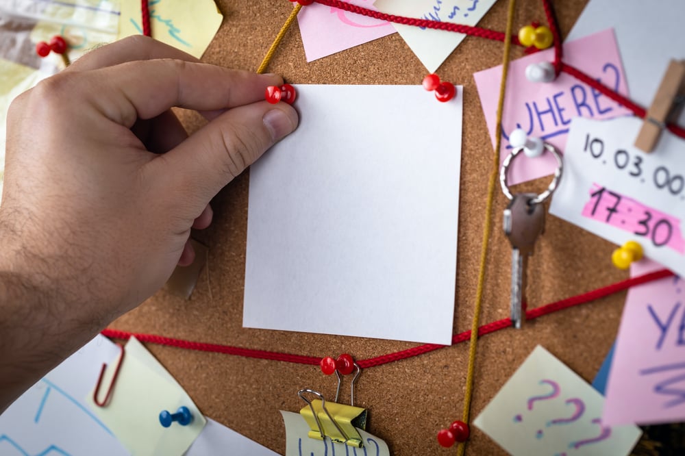 A hand pins a blank notecard to an investigation board loaded with sticky notes, pins, and red string.