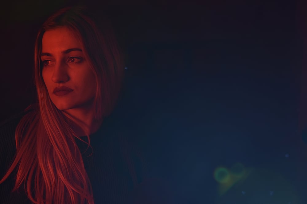 A person with long hair and red lipstick stands in the dark, looking off into the distance.