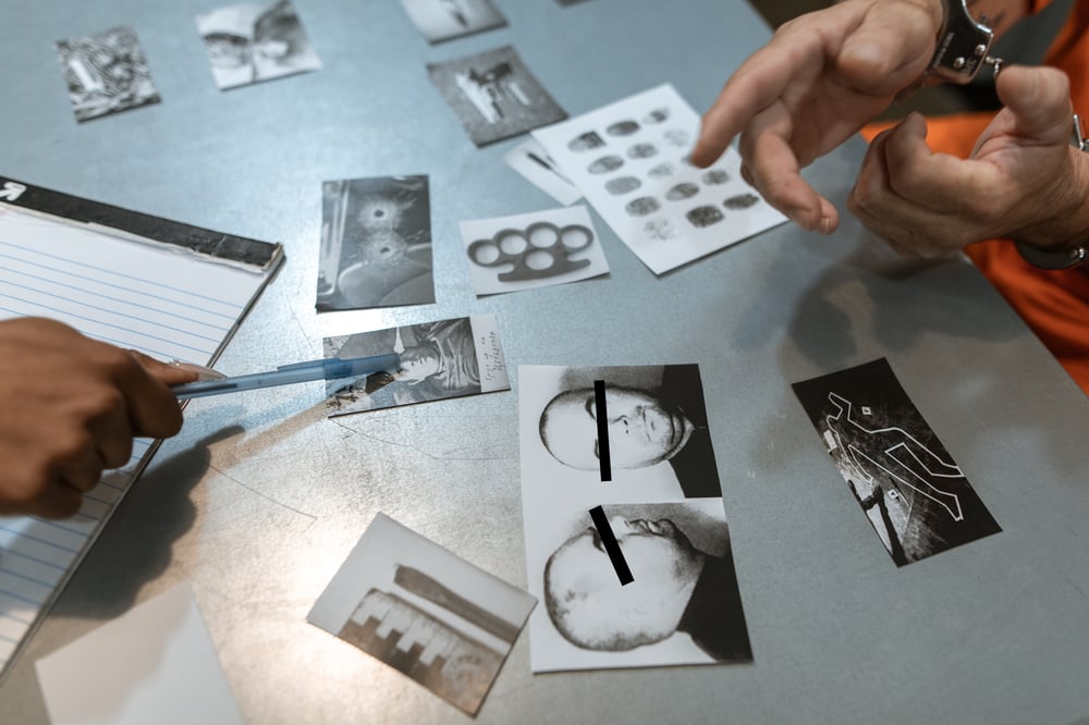 Two sets of hands—one handcuffed and one holding a pen—point to a set of evidence on a table.