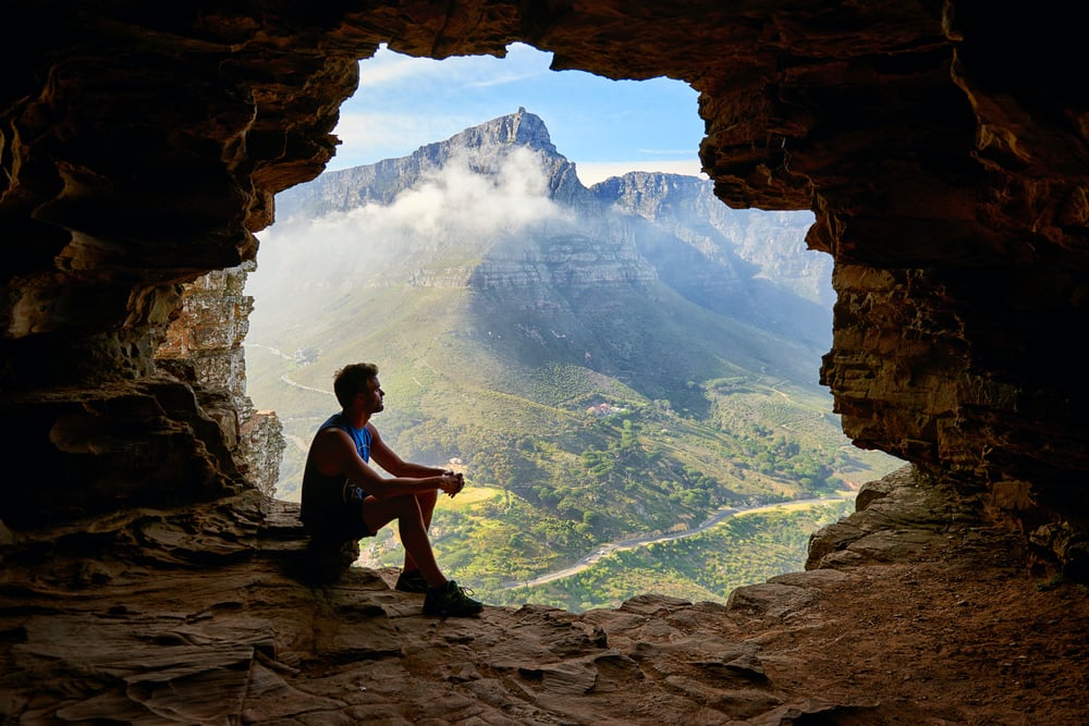 A person wearing a tank top and shorts sits in the opening of a cave, overlooking a green valley and gray mountains.