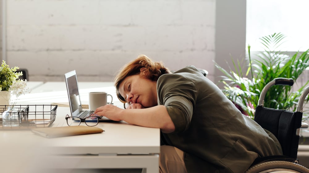 A person in a wheelchair asleep at a desk with their hand on the keyboard of a laptop.a