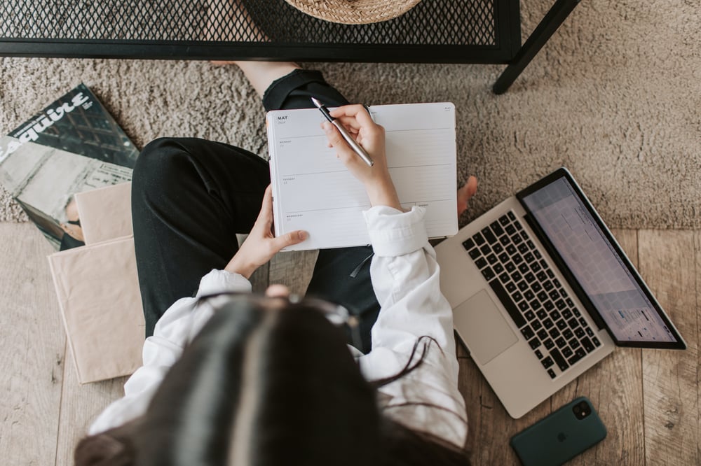 Overhead view of a person with long, dark hair sitting on a beige carpet and writing in a planner beside an open laptop. 