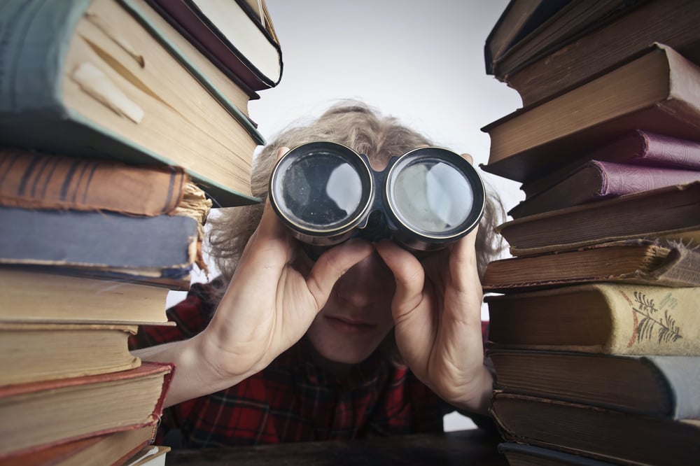 A person with blonde hair peering through binoculars between two stacks of old books.