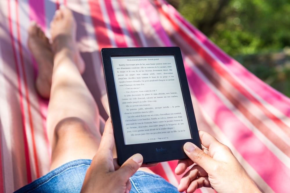 The subject's POV of their hands holding an e-reader and their legs stretched out in a pink and white striped hammock.