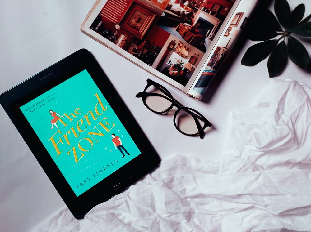 Overhead view of an e-reader displaying the book cover for The Friend Zone. Beside the e-reader is a pair of glasses, magazine, plant leaves, and a white bedsheet.