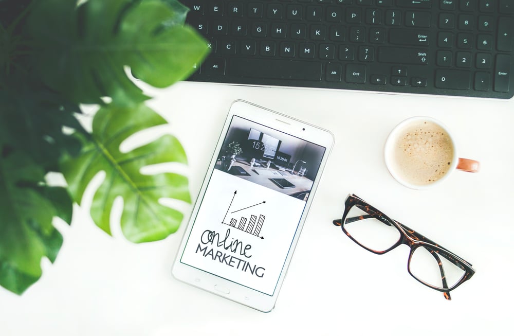 An overhead view of a white desk with glasses, a cup of coffee, a computer keyboard, a houseplant, and an e-reader with that says "online marketing" on the screen.