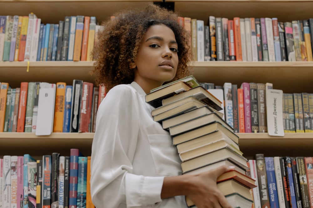 A person in a white shirt carrying a tall stack of books in front of full bookshelves.