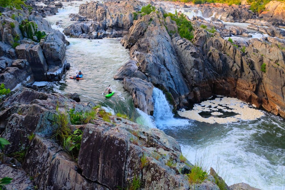 Three kayakers navigate kayaks along a the rapids of a canyon river, approaching a waterfall.