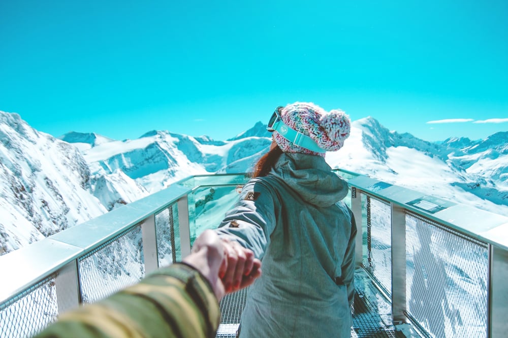A photo from the viewpoint of the photgrapher whose hand reaches out to hold the hand of another person. The other person stands in front, looking away from the camera and out over a snowy mountain range. They're wearing coat, knit hat, and ski goggles.