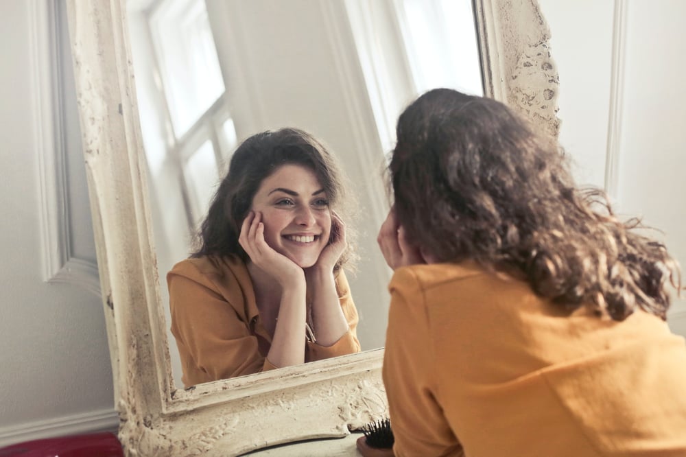 A person with long, wavy brown hair smiles at themselves in a shabby chic mirror.