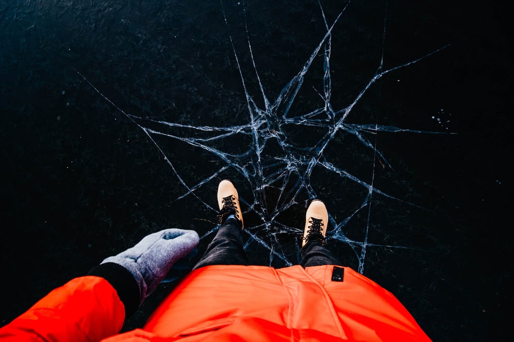 A first-person perspective looking down the front of the photographer's orange coat and at their tan boots standing on ice. A network of cracks is under their feet.