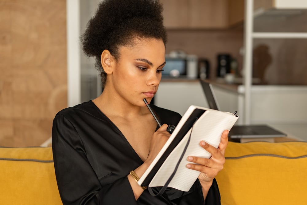 A person in a dark blouse taps their chin with a pen while looking thoughtfully at a notebook.