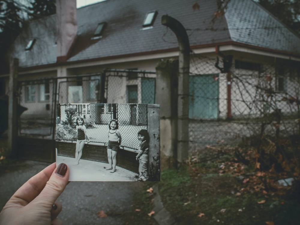 A hand holds and old black and white photo in front of a building. The photo shows children hanging out in the same spot years before.