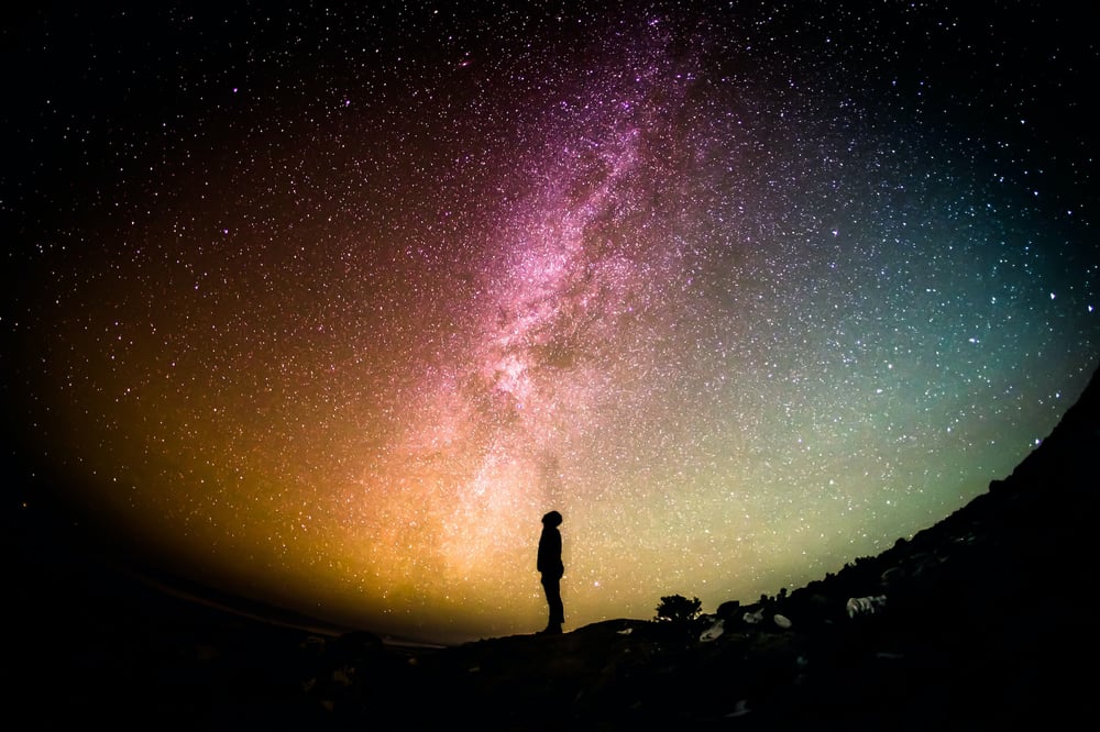 The silhouette of a person staring up at a multi-colored sky full of stars.