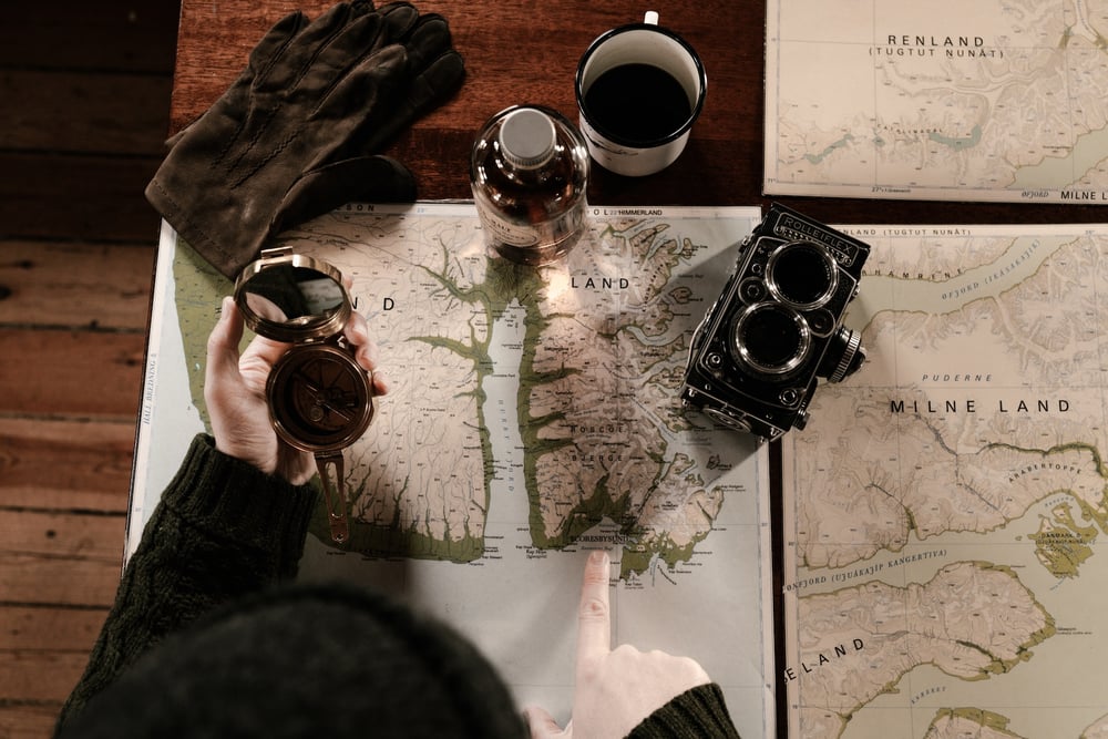 A person holds a compass and points to a map laying out on a desk beside other maps and a camera.