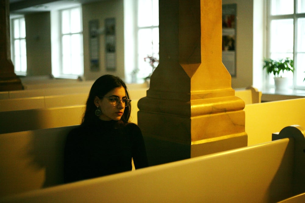 A person with long, dark hair and glasses sits alone in an empty church.