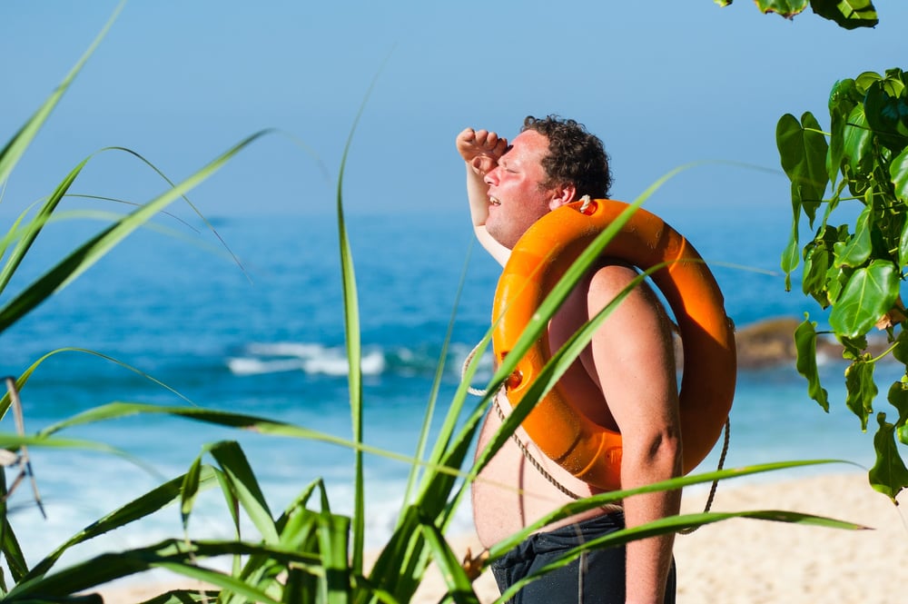 A person with life preserver looped around their arm stands on a beach wearing swim trunks and shielding their eyes with their hand as they search for something in the distance.